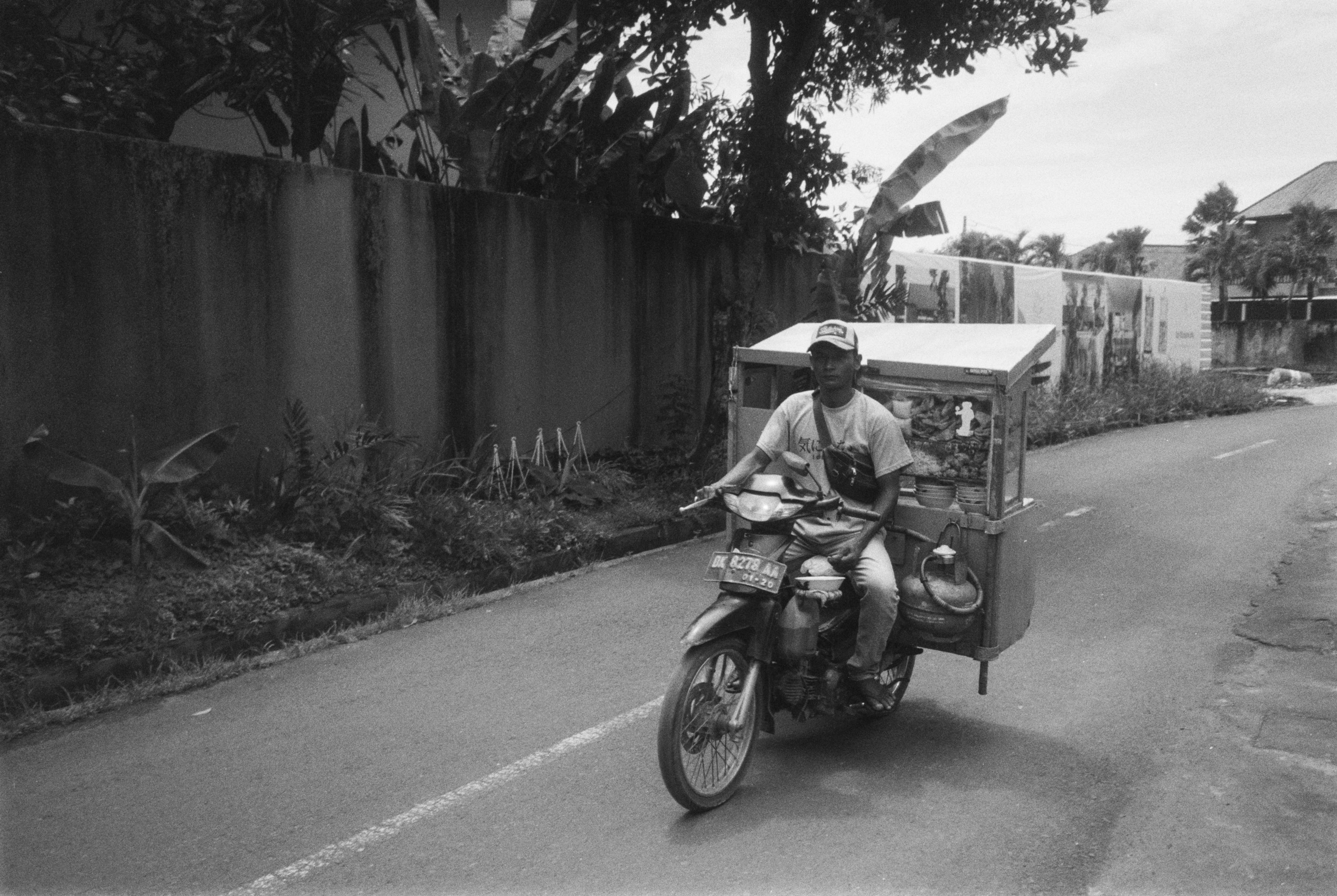 Black and white picture of a man driving a motorcycle carrying a food stand in the passenger's seat